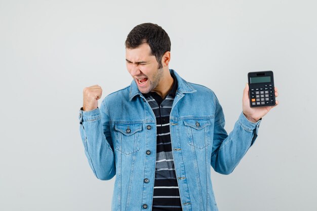 Jeune homme tenant la calculatrice, montrant le geste du gagnant en t-shirt, veste et à la chance.