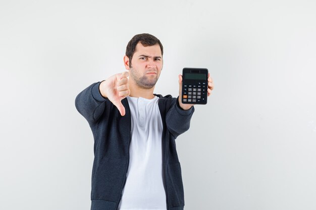 Jeune homme tenant la calculatrice et montrant le pouce vers le bas en t-shirt blanc et sweat à capuche noir zippé à l'avant et à la vue mécontent, de face.