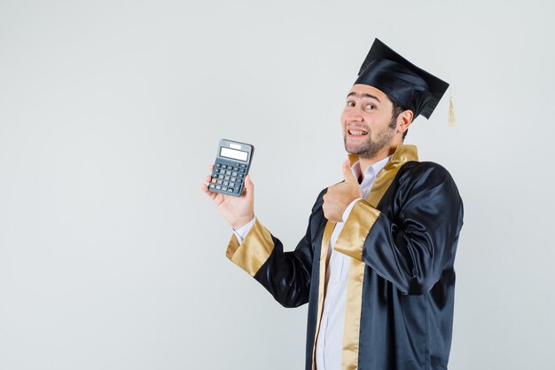 Jeune homme tenant la calculatrice, montrant le pouce vers le haut en uniforme d'études supérieures et à la joyeuse vue de face.