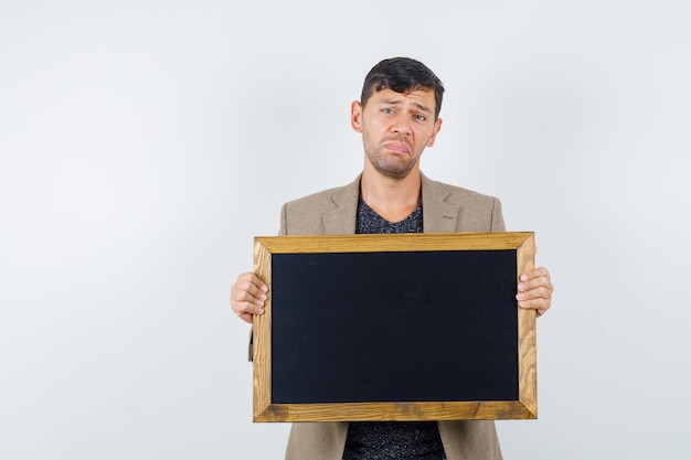 Jeune homme tenant un carton en veste marron grisâtre, chemise noire et à la triste. vue de face.