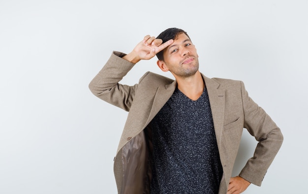 Jeune homme tenant le doigt sur son front en veste marron grisâtre, chemise noire et à la bizarre. vue de face.