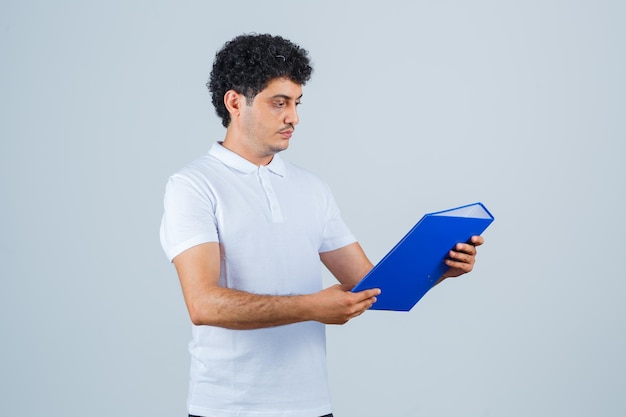 Jeune homme tenant un dossier et le regardant en t-shirt blanc et jeans et regardant concentré, vue de face.