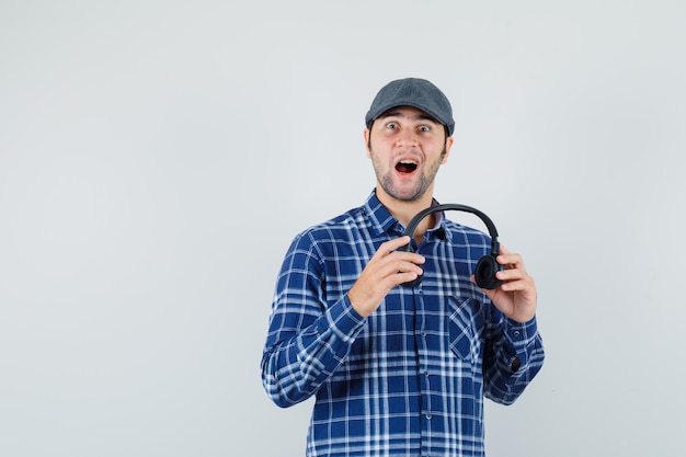 Jeune homme tenant des écouteurs en chemise, casquette et à la surprise. vue de face.