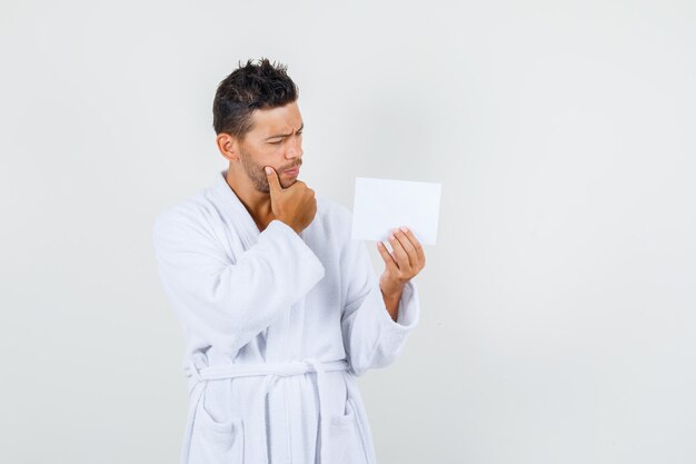 Jeune homme tenant une feuille de papier avec la main sur le menton en peignoir blanc et à la pensif. vue de face.