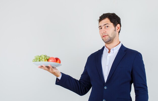 Jeune homme tenant des fruits en assiette en costume et à la confiance. vue de face.