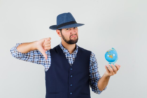 Jeune homme tenant un globe, montrant le pouce vers le bas en chemise, gilet, chapeau et à la recherche de mécontentement, vue de face.