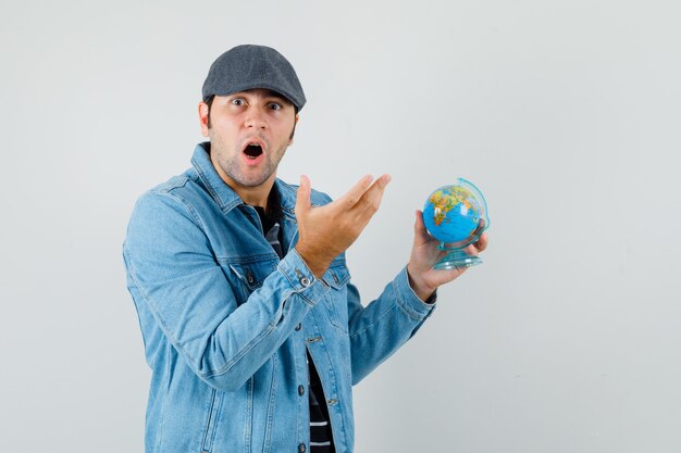 Jeune homme tenant un globe tout en regardant la caméra en veste, casquette et à la surprise