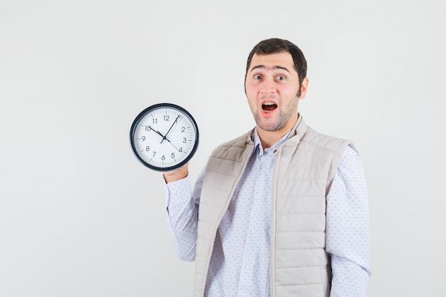 Jeune homme tenant une horloge dans une main en veste beige et casquette et à la surprise. vue de face.