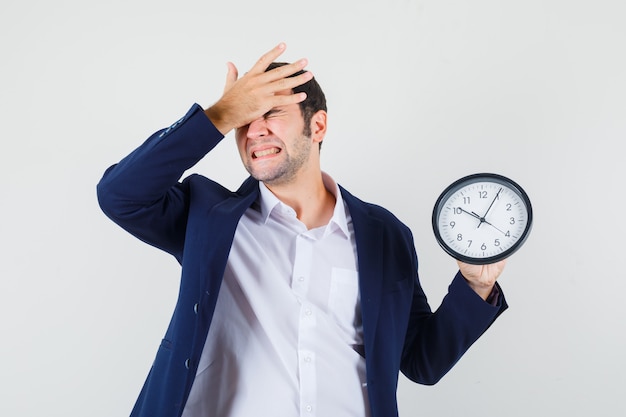 Photo gratuite jeune homme tenant une horloge murale avec la main sur le front en chemise et veste et à oublier