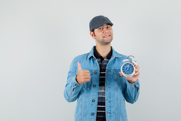 Jeune homme tenant une horloge tout en montrant le pouce vers le haut en veste, casquette et à la satisfaction.