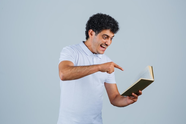 Jeune homme tenant un livre et le pointant en t-shirt blanc et jeans et l'air heureux, vue de face.