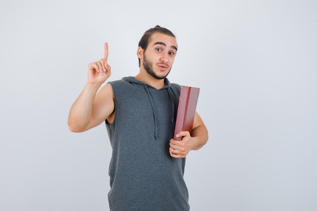 Jeune homme tenant un livre tout en montrant pointant vers le haut en sweat à capuche sans manches et à la vue de face, confiant.