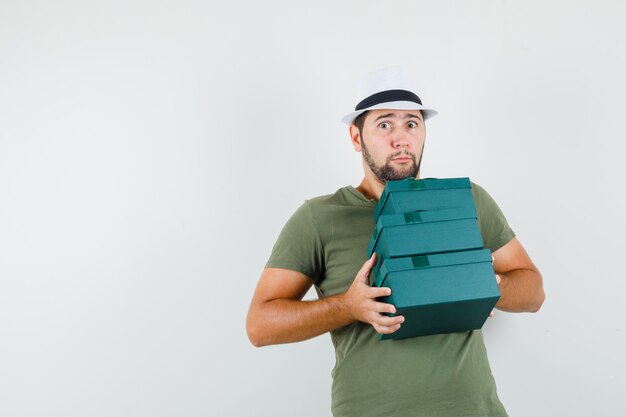 Jeune homme tenant de lourdes boîtes présentes en t-shirt vert et chapeau et à la perplexité