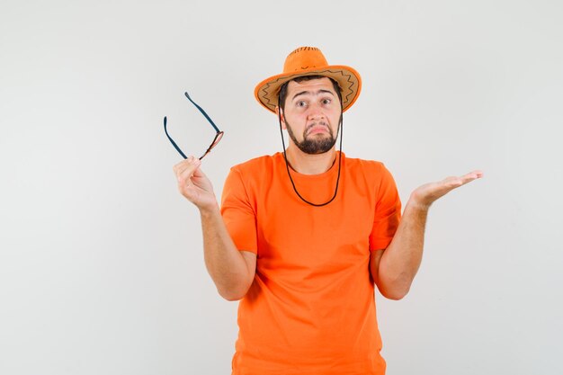 Jeune homme tenant des lunettes avec un geste impuissant en t-shirt orange, chapeau, vue de face.