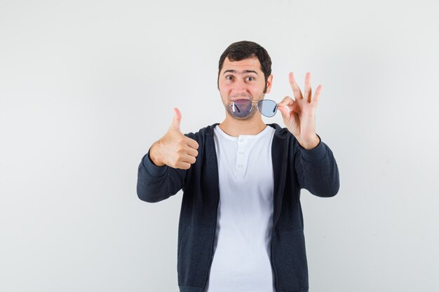 Jeune homme tenant des lunettes, montrant le pouce vers le haut en t-shirt, veste et à la confiance. vue de face.