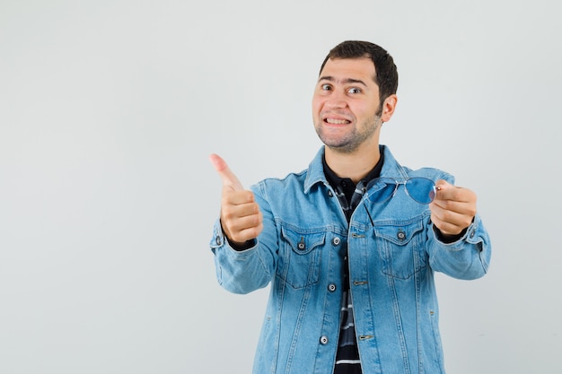 Jeune homme tenant des lunettes, montrant le pouce vers le haut en t-shirt, veste et à la satisfaction