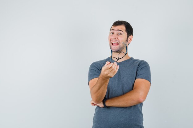 Jeune homme tenant des lunettes en t-shirt gris et à la recherche de plaisir. vue de face.