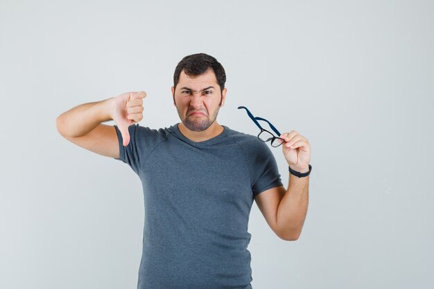 Jeune homme tenant des lunettes en t-shirt gris et regardant mécontent, vue de face.