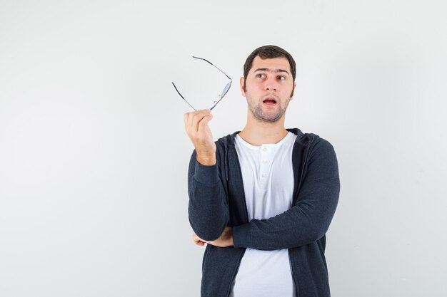 Jeune homme tenant des lunettes en t-shirt, veste et à la réflexion. vue de face.