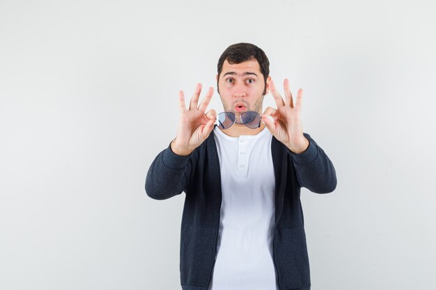 Jeune homme tenant des lunettes en t-shirt, veste et regardant étonné, vue de face.