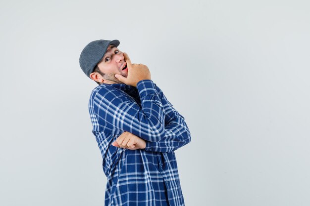 Jeune homme tenant la main sur la bouche en chemise, casquette et à la recherche d'optimiste. vue de face.