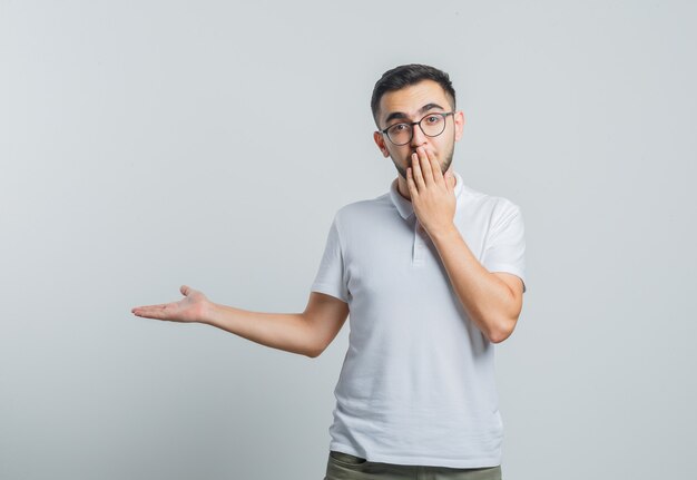 Jeune homme tenant la main sur la bouche, montrant quelque chose en t-shirt blanc, pantalon et regardant concerné