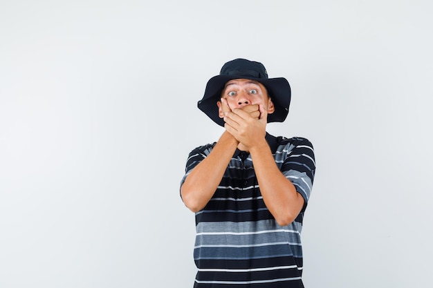 Jeune homme tenant la main sur la bouche en t-shirt, chapeau et ayant l'air effrayé. vue de face.