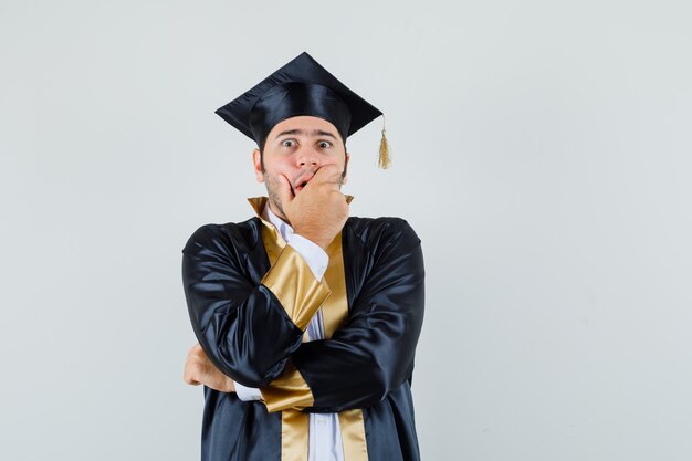 Jeune homme tenant la main sur la bouche en uniforme d'études supérieures et l'air alarmé. vue de face.