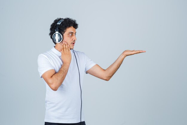 Jeune homme tenant la main sur la joue, écartant la paume en t-shirt blanc et l'air choqué. vue de face.