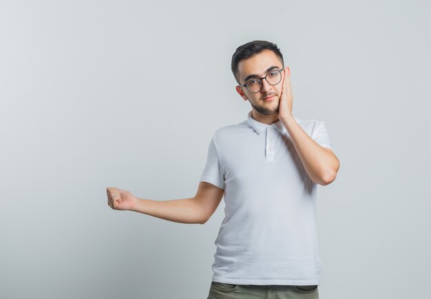 Jeune homme tenant la main sur la joue, pointant vers l'extérieur en t-shirt blanc, pantalon et à sensible