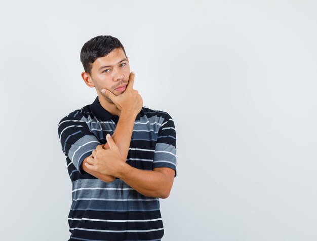 Jeune homme tenant la main sur le menton en t-shirt et semblant sensible, vue de face.