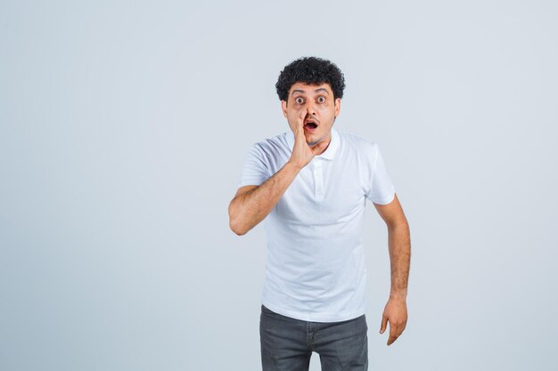 Jeune homme tenant la main près de la bouche comme appelant quelqu'un en t-shirt blanc et jeans et l'air surpris, vue de face.