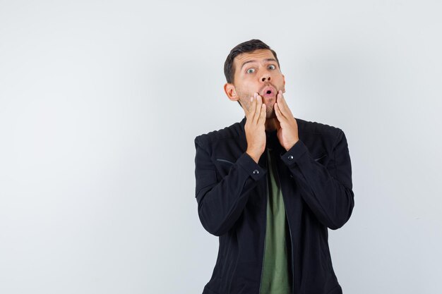 Jeune homme tenant la main près de la bouche ouverte en t-shirt, veste et se demandait. vue de face.