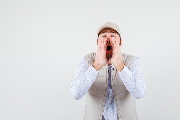 Jeune homme tenant la main près de la bouche tout en criant en veste beige et casquette et à la recherche de sérieux. vue de face.