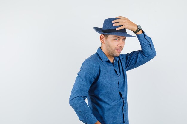 Jeune homme tenant la main sur son chapeau en chemise bleue et regardant gai. vue de face.