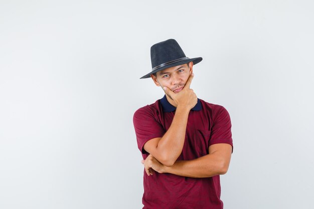 Jeune homme tenant la main sur son menton en chemise rouge, chapeau noir et à la vue réfléchie, de face.