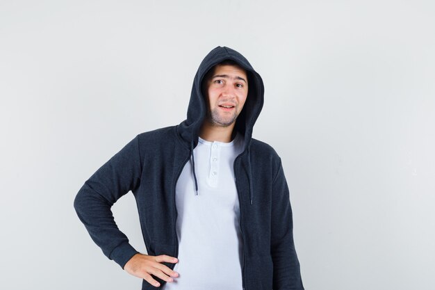 Jeune homme tenant la main sur la taille en t-shirt, veste et à la confiance. vue de face.