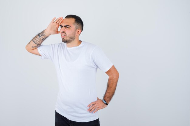 Jeune homme tenant la main sur la taille tout en mettant la main près du visage en t-shirt blanc et pantalon noir et l'air pensif