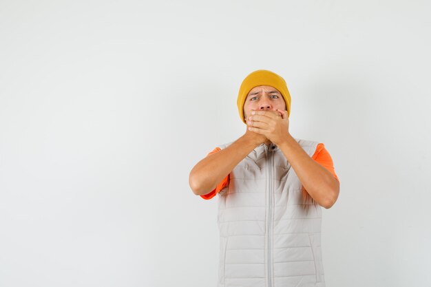 Jeune homme tenant les mains sur la bouche en t-shirt, veste, chapeau et à la peur, vue de face.