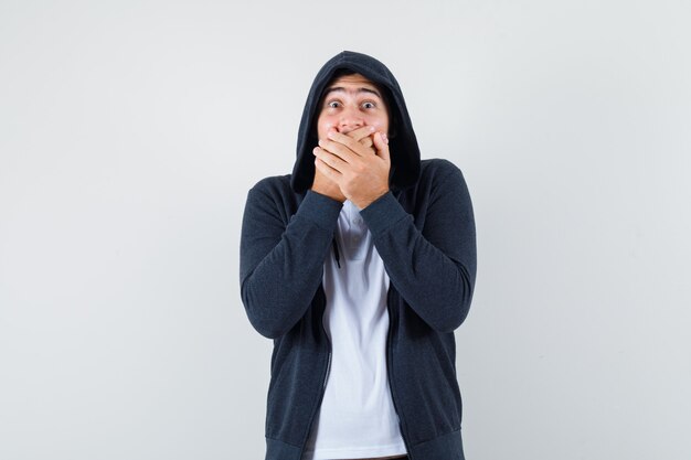 Jeune homme tenant les mains sur la bouche en t-shirt, veste et à la vue étonnée, de face.