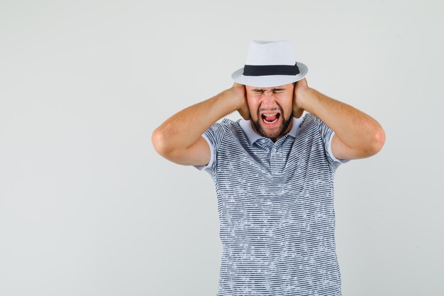 Jeune homme tenant les mains sur les oreilles en t-shirt, chapeau et l'air irrité. vue de face.