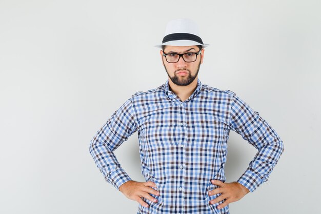 Jeune homme tenant les mains sur la taille en chemise à carreaux, chapeau et à la vue irrésolue, de face.