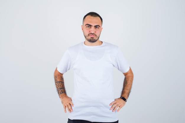 Jeune homme tenant les mains sur la taille en t-shirt blanc et pantalon noir et à la sérieuse