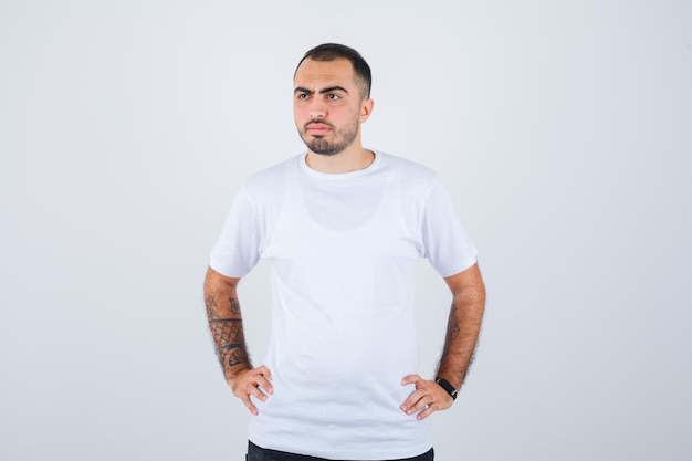 Jeune homme tenant les mains sur la taille en t-shirt blanc et pantalon noir et à la sérieuse