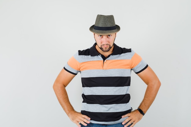 Jeune homme tenant les mains sur la taille en t-shirt, chapeau et à la vue de face stricte.