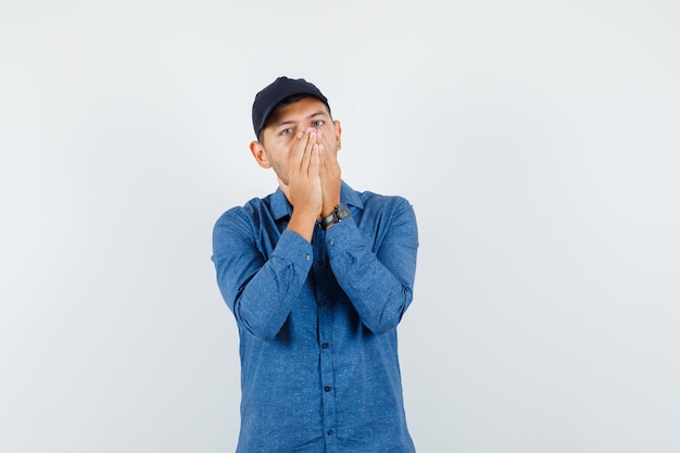 Jeune homme tenant les mains sur le visage en chemise bleue, casquette et à la peur. vue de face.