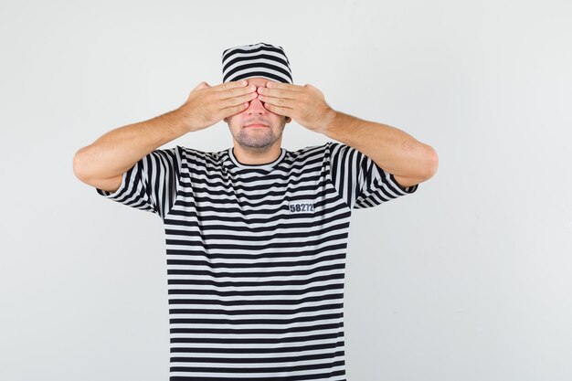 Jeune homme tenant les mains sur les yeux en t-shirt, chapeau et regardant calme, vue de face.