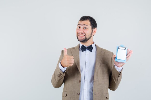 Jeune homme tenant un mini presse-papiers avec le pouce vers le haut en costume et à la bonne humeur. vue de face.