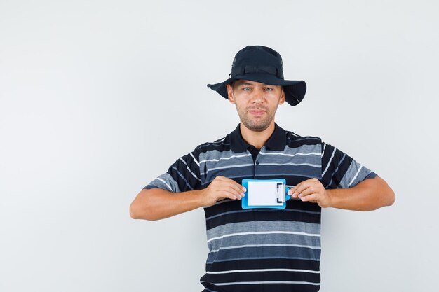 Photo gratuite jeune homme tenant un mini presse-papiers en t-shirt, chapeau et regardant positif, vue de face.