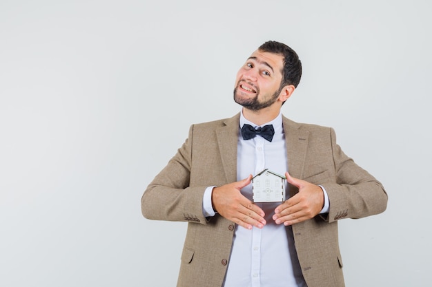 Photo gratuite jeune homme tenant un modèle de maison en costume et à la recherche de bonne humeur. vue de face.